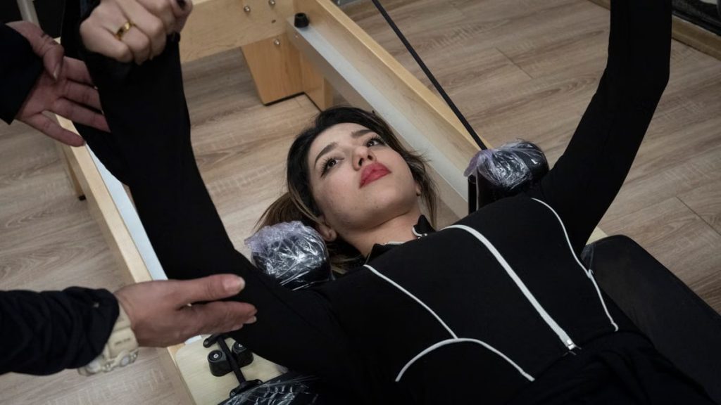 Woman in black outfit performing Pilates exercise with assistance in gym.